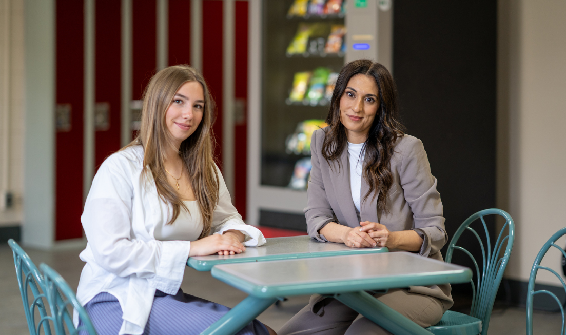 Anthea Christoforou and Angelina Baric sitting at a food-court-style table with a vending machine in the background.