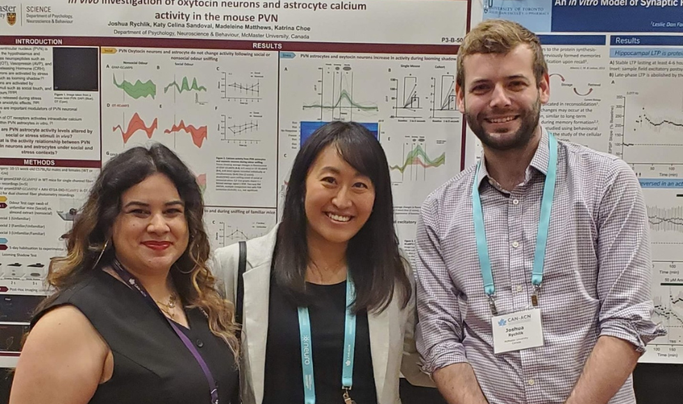 Three people smile and pose for the camera in front of a board covered in complex graphs.