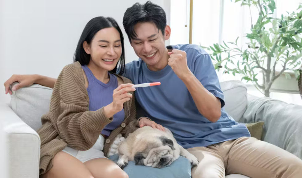A couple sit on a couch with their dog, looking at a pregnancy test with smiles.