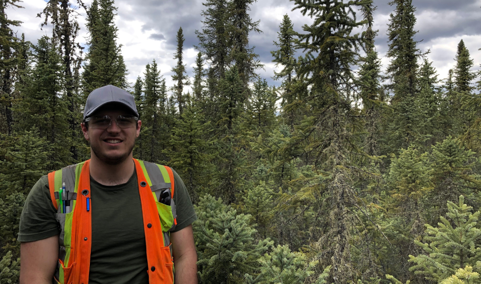 A man in a safety vest stands in front of a forest.