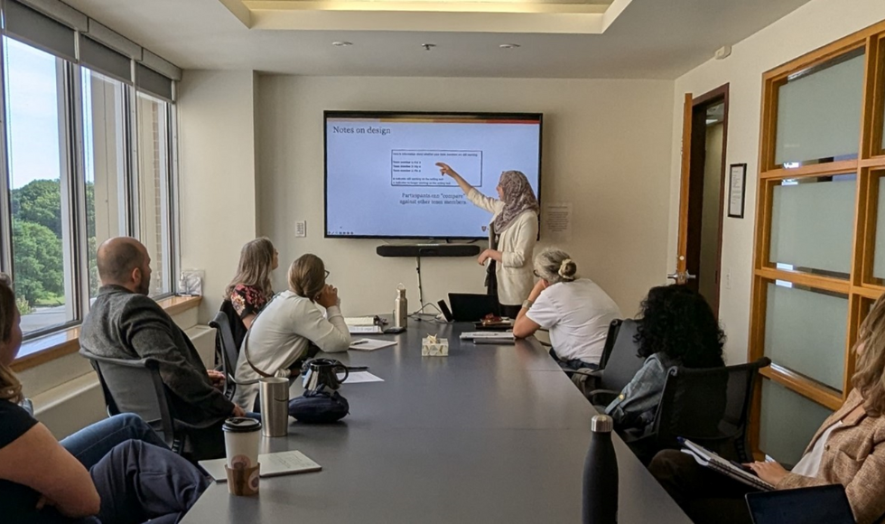 Several people sit around a large meeting table, all looking at a screen on the wall, with one person pointing to something on the screen.