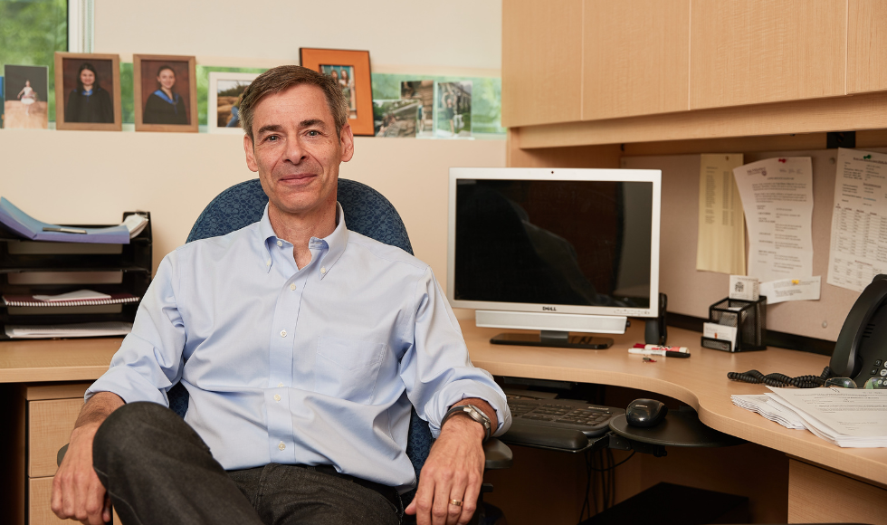 A man sits at a desk, facing the camera. Behind him there is a computer on the desk.