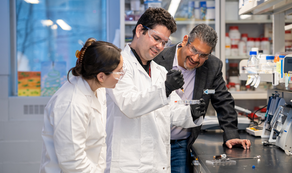 Two people with safety goggles in a lab stand on either side of a third person, watching him pippette something into a test tube.