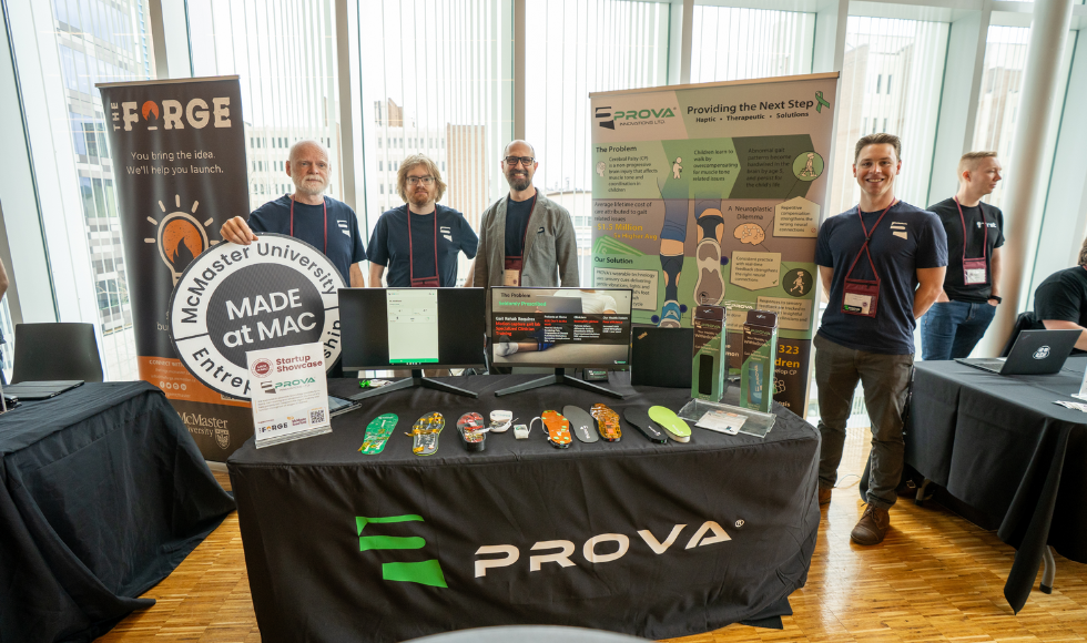 Four men stand behind a table, which has a tablecloth on it that reads 'PROVA' and has several shoe insoles sitting on it, along with computer screens. One man is holding a sign that reads 'Made at Mac, McMaster University.' The edge of two other tables are visible on the left and right, and another man is standing behind the table to the right.