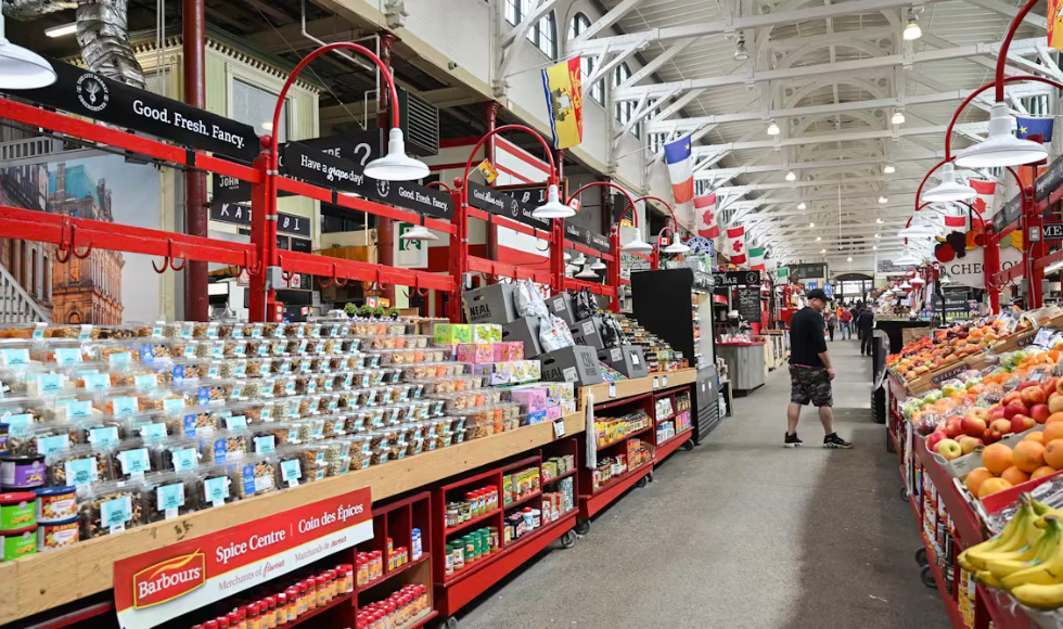 An aisle of a grocery store, with containers of nuts and spices on one side of the aisle, and fresh fruit including bananas, apples and oranges on the other side of the aisle. A man stands partway down the aisle, looking towards the fruit.