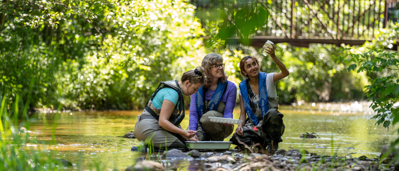 Three women kneel on the edge of a river. They are wearing vests and hip waders, holding collection equipment. One of them holds a jar of water up and looks at it.
