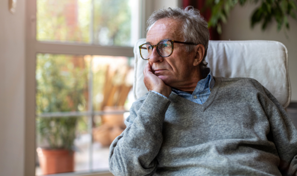 An older man sits and stares out a window, looking pensive.