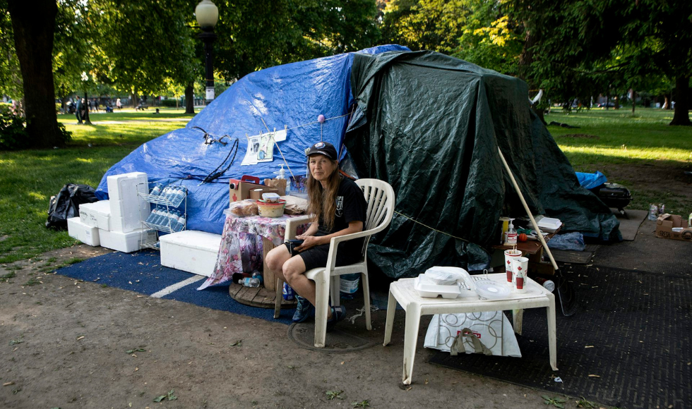 A woman sits outside on a plastic chair in front of a structure made of tarps in a city park. A table with coffee, a shelf with water bottles, and styrofoam blocks are beside her.