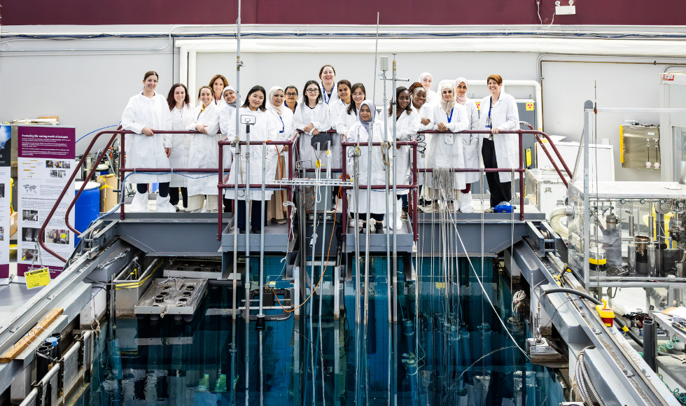 A group of women in lab coats stand on a metal terrace above a lab.