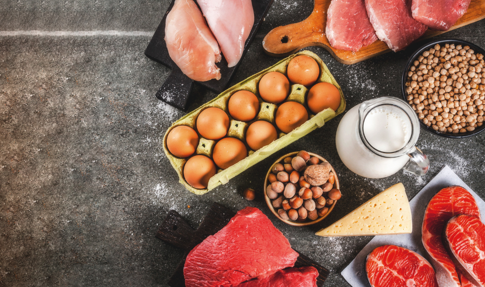 Several different types of protein-rich foods sit on a dark table, including eggs, cheese, milk, nuts, and meat.