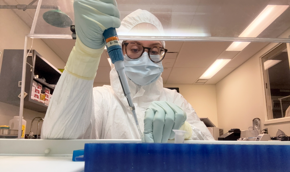 In a lab, a woman in a full cleanroom suit, mask, and gloves holds a dropper above a tray.