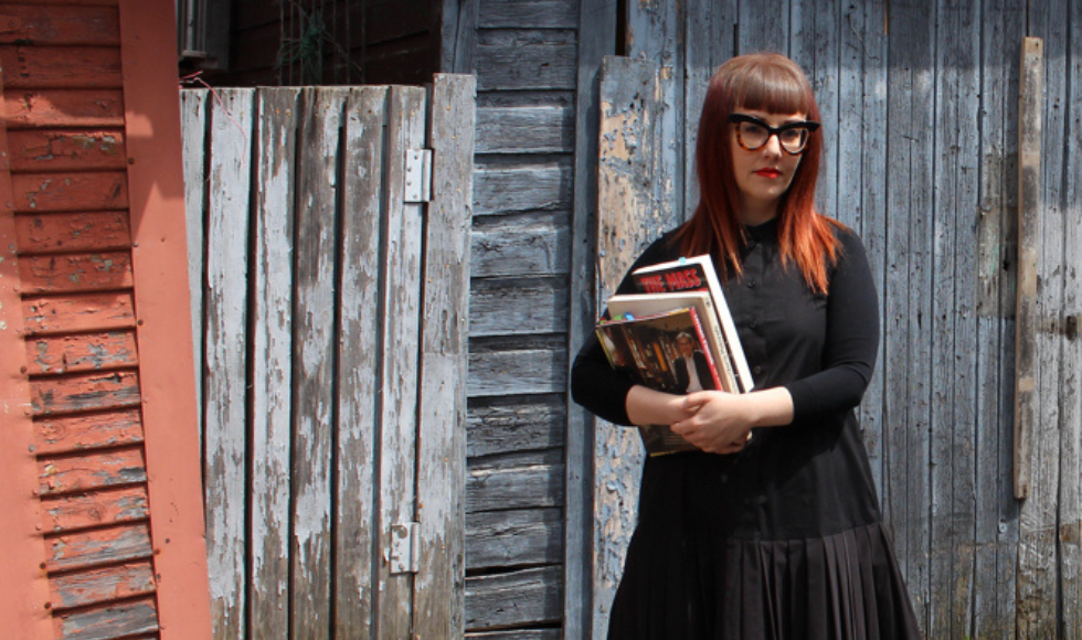 A woman stands outside of a barn wall, holding several books in her arms.