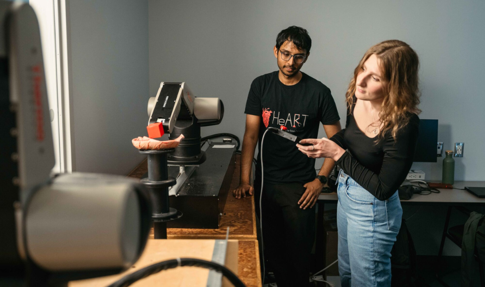 Two young people stand at a wooden counter, looking at a robotic arm sitting on the counter.