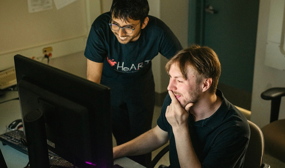 Two young men look at a laptop screen, smiling.