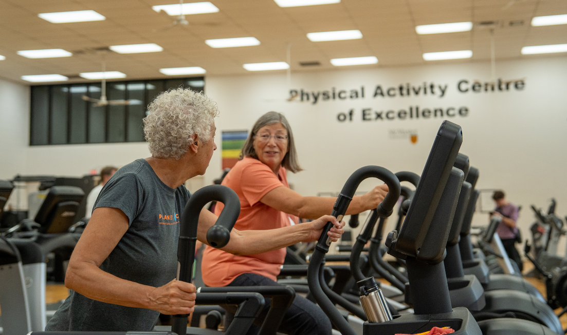 Two smiling older adults working out in an indoor gym with the worlds Physical Activity Centre of Excellence on the wall behind them.