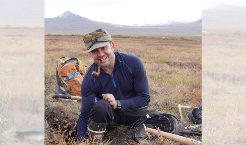 A man kneels on the ground in a large field, with mountains far in the distance behind him. He is holding a pipe in his mouth, and various equipment - including a backpack, shovel, and cord - are lying on the ground around him.