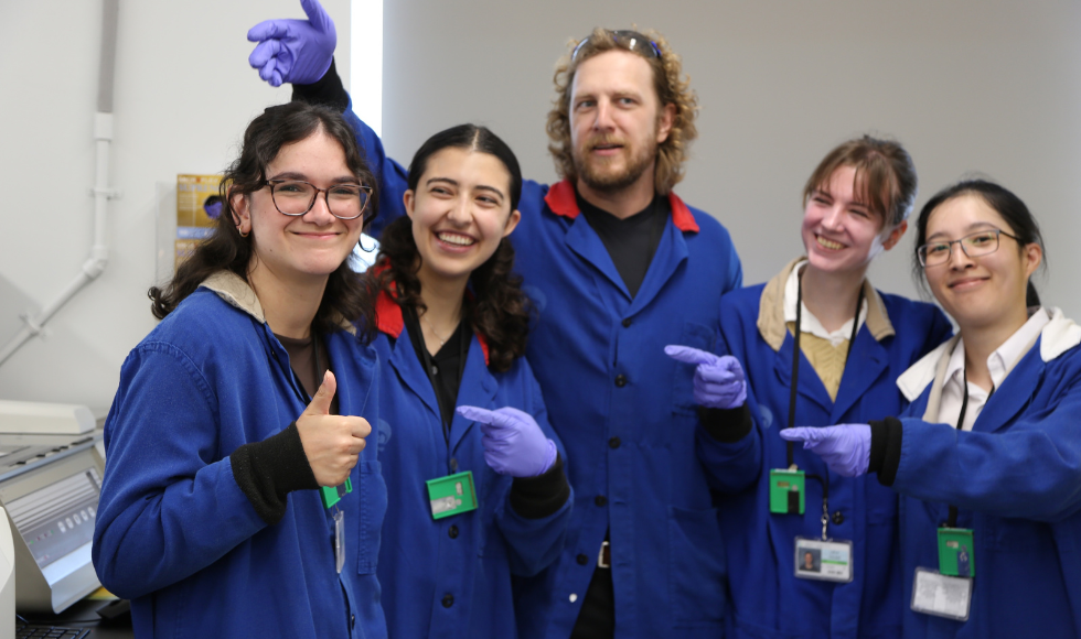 Five people in blue jumpsuits stand in a lab. Four of them are pointing to the woman on the far left, who is smiling and giving a thumbs-up.