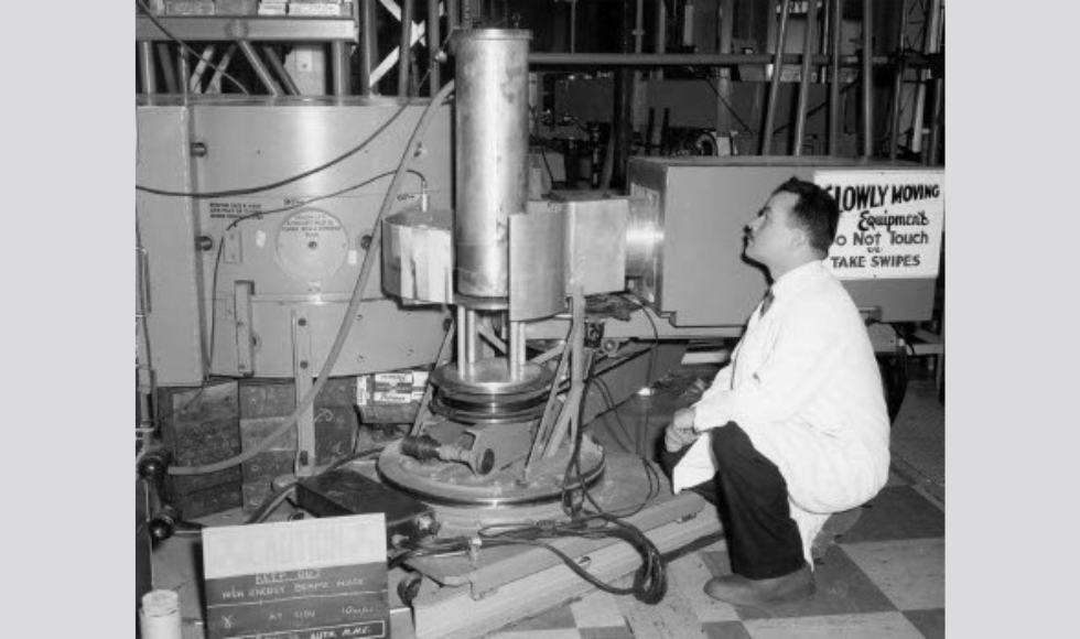 A black and white photo of a man in a lab coat, crouching beside a machine.