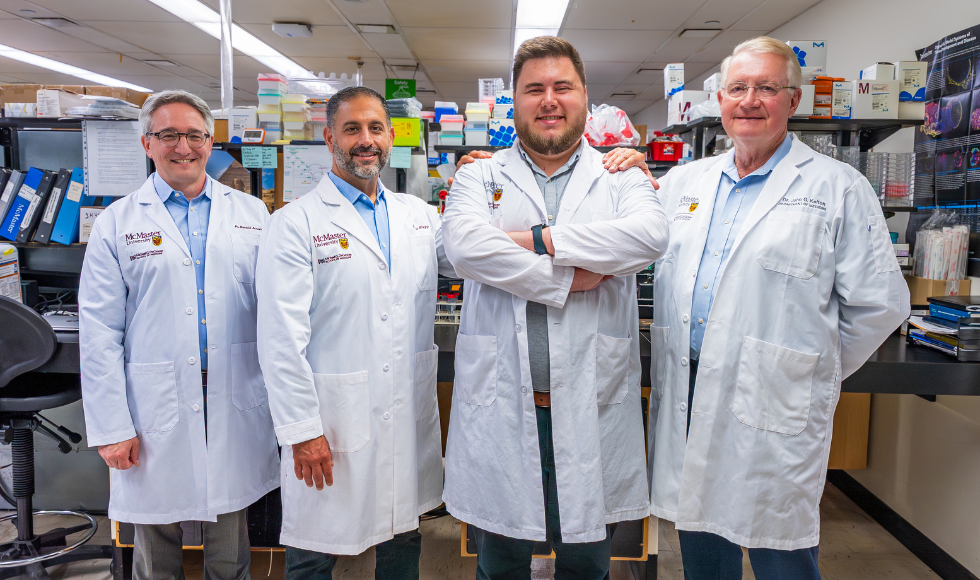 Four scientists stand in white lab coats shoulder to shoulder in a lab.