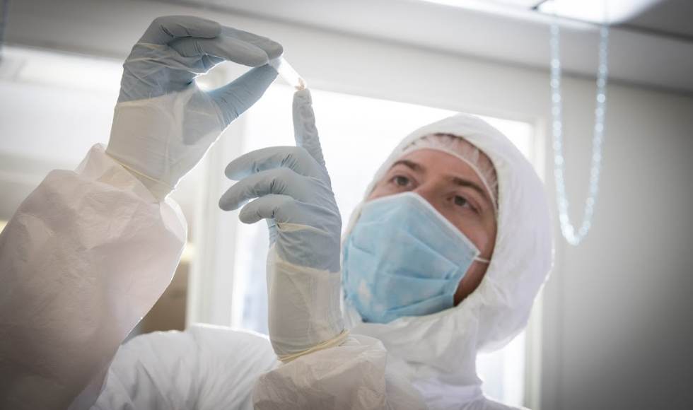 A person in a white lab suit, mask, and gloves holds up a small vial, looking at it.